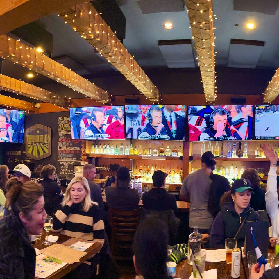 Patrons at The Fields Bar & Grill watching sports on multiple screens above a liquor bar.