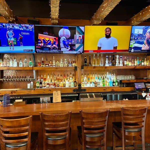 A well-stocked bar at The Fields Bar & Grill with multiple TVs showing sports above.