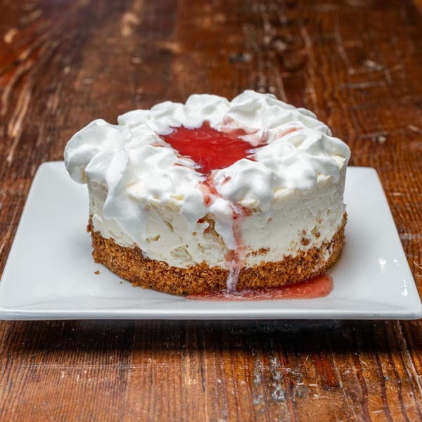 Cheesecake with a graham cracker crust, topped with whipped cream and strawberry sauce on a white plate.